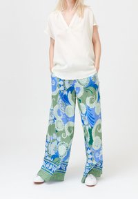 Kvinna som bär en lös vit blus med kort ärm, vida byxor med blå och gröna blommiga paisleymönster, och vita sneakers, med händerna i fickorna.