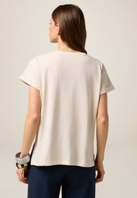 Tricou bej cu mânecă scurtă, cu croială lejeră, decolteu rotund și fente laterale, asortat cu pantaloni închiși la culoare și brățări suprapuse.