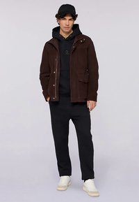 Hnědá semišová bunda přes černou mikinu s kapucí, doplněná černými kalhotami a krémovými teniskami. Model má na hlavě černou čepici. Ležérní outfit s vrstveným vzhledem.