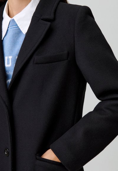 Cappotto di lana nero con revers strutturati, taschino sul petto e una vestibilità rilassata. Il colletto superiore si abbina a una felpa azzurro chiaro sottostante.