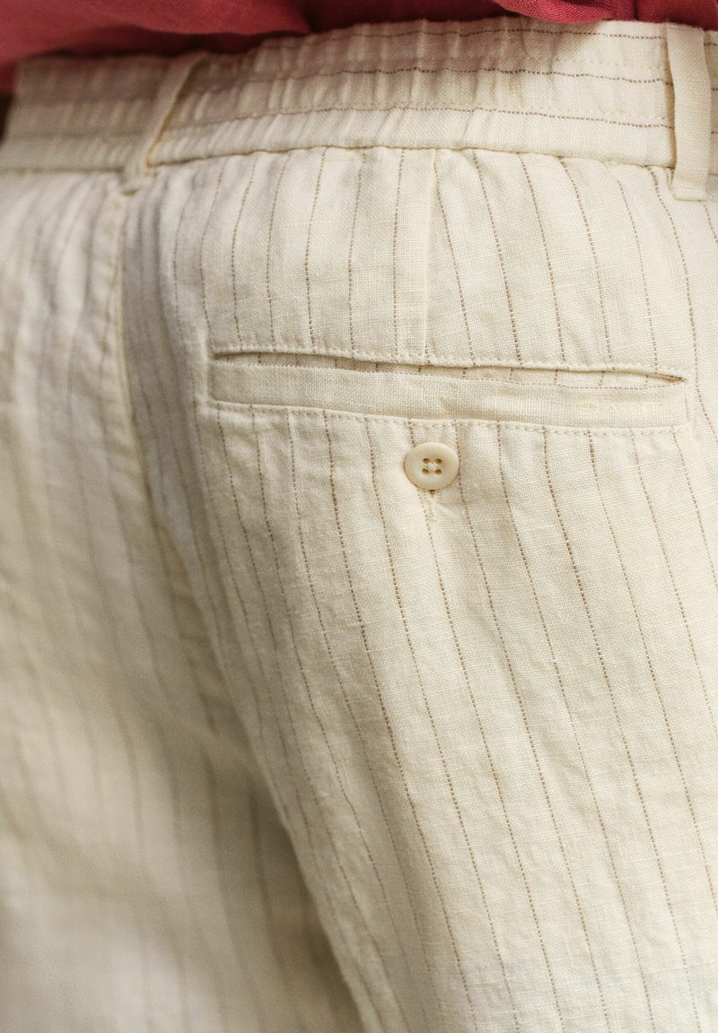 Primer plano del bolsillo trasero de pantalones beige a rayas con cierre de botón y trabillas para cinturón, usados con cinturilla de tela roja visible.