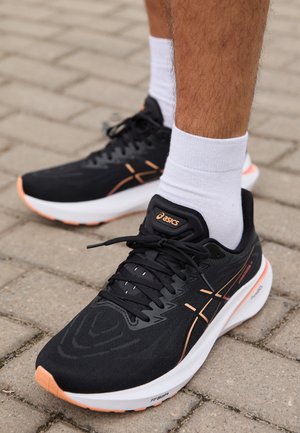 Black running shoes with a textured mesh upper, featuring orange accents, a white midsole, and black laces, positioned on a paved surface.