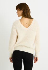 Suéter de cuello en V en un tejido crema suave y esponjoso con un ajuste relajado. Cuenta con hombros caídos y dobladillo acanalado. Combinado con jeans ajustados negros.