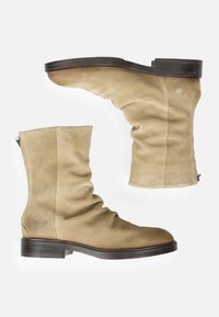 Botas de tobillo de ante beige con un diseño arrugado, suela de goma marrón plana y logo de mariposa decorativo en el talón. Cierre de cremallera en la parte posterior.