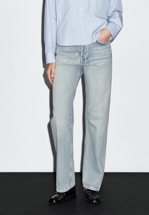 Persoon die lichtblauwe jeans met rechte pijpen, een lichtblauw overhemd en zwarte loafers draagt, staand met één hand in de zak.