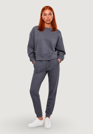Ung kvinde med rødt hår iført en matchende mørkegrå sweatshirt og joggingbukser med hvide sneakers, stående med hænderne i lommerne.