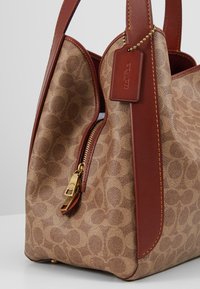 Sac à main marron à motifs avec poignées en cuir, fermeture éclair dorée et étiquette en cuir attachée sur une surface blanche.