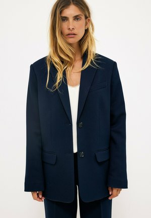 Frau mit langen blonden Haaren, die einen marineblauen übergroßen Blazer und ein weißes Oberteil vor einem einfarbigen weißen Hintergrund trägt.