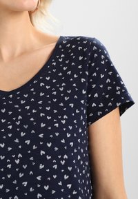 Camiseta de cuello en V azul marino hecha de tela suave, con un patrón de corazones plateados dispersos y mangas cortas.