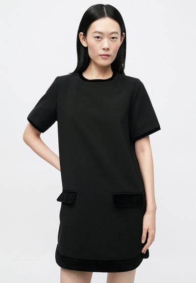 Mujer de pie con un vestido negro de manga corta, con dos bolsillos delanteros con solapa y un escote redondo, sobre un fondo liso.