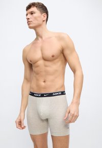 Μυώδης άνδρας όρθιος, χωρίς μπλούζα, φοράει ανοιχτό γκρι μπόξερ της Nike με μαύρη ζώνη μέσης, κοιτάζοντας αριστερά σε απλό φωτεινό φόντο.