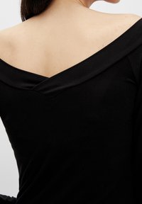 Frau trägt ein schwarzes Oberteil mit schulterfreiem Schnitt, zeigt ihren oberen Rücken und hat eine entspannte Haltung vor einem schlichten Hintergrund.