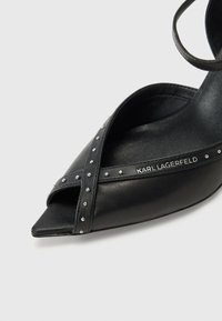 Zwarte leren sandalen met hak en een spitse neus, voorzien van gekruiste enkelbandjes en versierd met zilveren studs. Het 'KARL LAGERFELD' logo is zichtbaar.