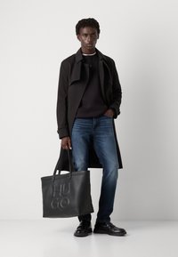 Bolso tote de cuero negro con el logo "HUGO" en relieve, llevado por un modelo masculino con un abrigo trinchera oscuro, suéter negro y jeans azules.