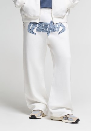 Persona che indossa pantaloni bianchi a gamba larga con scritta a patch in denim blu "faster" in vita e sneakers beige con dettagli in mesh.