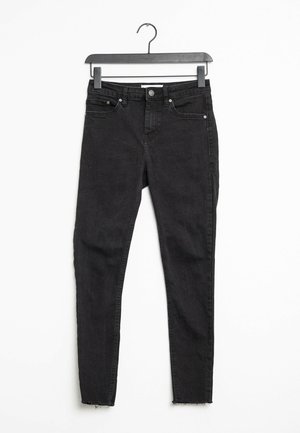 Jean skinny noir taille haute avec cinq poches et fermeture à bouton, suspendu sur un cintre noir contre un fond blanc.
