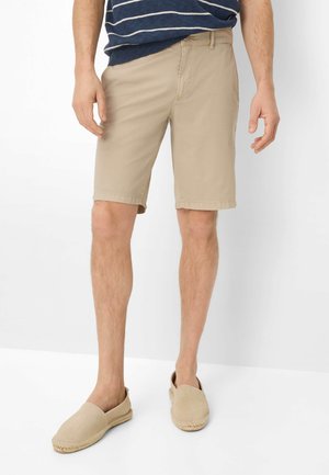 Mand iført beige knælange shorts, beige slip-on sko og en navyblå stribet skjorte, stående mod en hvid baggrund.