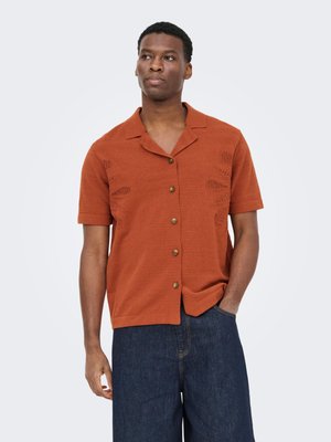 Homme portant une chemise orange brûlé à manches courtes avec des motifs texturés et un jean large bleu foncé, debout avec une main dans la poche.