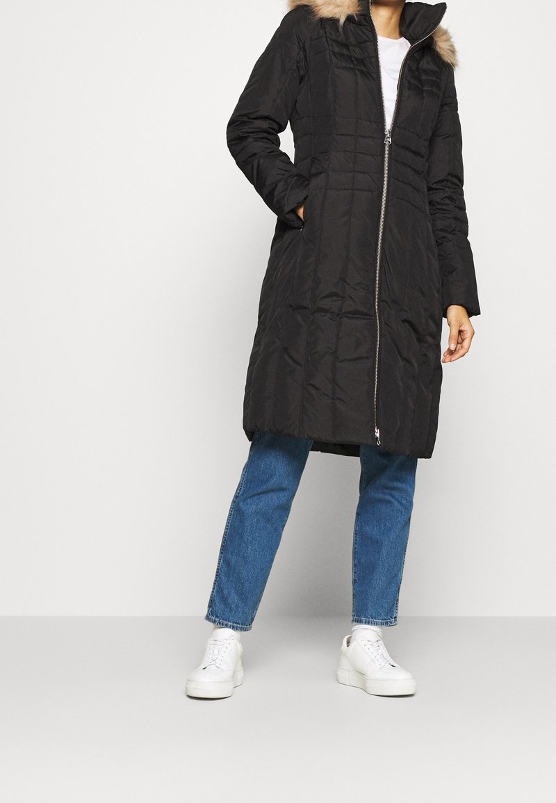 Svart puff-jacka med dragkedja fram, kviltat mönster och huva med fuskpälsfoder. Kombinerad med blå jeans och vita sneakers.