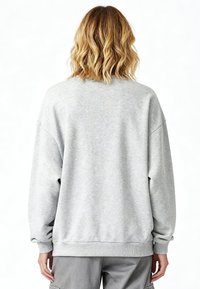Personne aux cheveux blonds ondulés mi-longs portant un sweat-shirt ample gris clair et un pantalon gris, vue de dos sur un fond blanc.