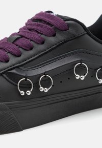 Baskets noires avec des lacets violets, présentant des anneaux métalliques avec des perles sur le côté. Surface en cuir lisse et semelle en caoutchouc avec une adhérence texturée.
