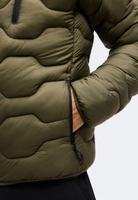 Homme portant une veste matelassée vert olive avec une main dans la poche zippée latérale sur un fond uni.