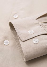 Gros plan sur le poignet et la manche beige d'une chemise avec des boutons blancs et des détails de couture visibles.
