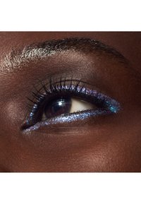 Primo piano di un occhio di pelle scura con lunghe ciglia e eyeliner glitter iridescente blu-viola sulle palpebre superiore e inferiore.