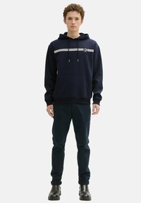 Sudadera navy con rayas blancas en el pecho, bolsillo tipo canguro y capucha con cordón, combinada con pantalones oscuros y zapatos negros.
