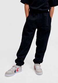 Sorte joggingbukser med sidelommer, justerbar talje og struktureret stof. Kombineret med grå sneakers med røde og marineblå detaljer.