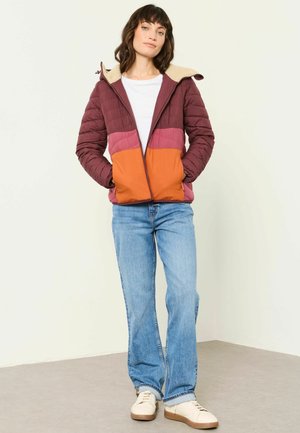 Frau steht mit Händen in den Taschen und trägt eine farblich abgestimmte Steppjacke in Bordeaux, Rosa und Orange, blaue Jeans und weiße Sneaker.