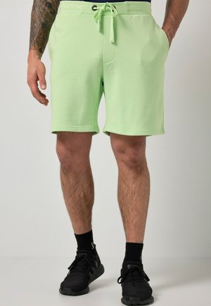 Mężczyzna w jasnooliwkowych szortach ze ściągaczem, czarnych skarpetkach i czarnych trampkach, stoi z rękami częściowo w kieszeniach.