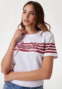 Witte katoenen T-shirt met rode horizontale strepen en een touw geïnspireerd grafisch ontwerp op de borst. Korte mouwen en een ronde hals.