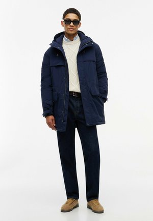Mann trägt dunkelblaue Parka, weißen Zopfpullover, dunkle Jeans, beige Sneakers und schwarze Sonnenbrille und steht vor weißem Hintergrund.