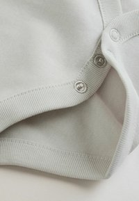 Šviesiai pilka medvilninė medžiaga su lygiu paviršiumi, krašte su spaudžiamosiomis sagomis ir ribbinku megztu apačios kraštu papildomam akcentui.