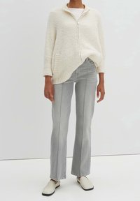 Kermanvärinen vetoketjullinen neulepaita, jossa on teksturoitu neulos ja sivutaskut, yhdistetty vaaleanharmaisiin suoriin farkkuihin ja kermanvärisiin slip-on-kenkiin.