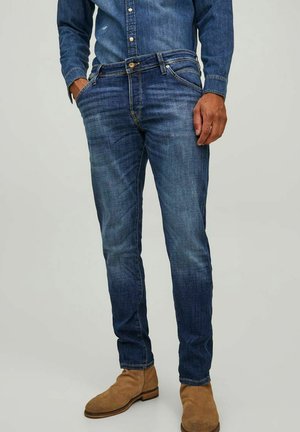 Jeans de mezclilla azul oscuro con un corte slim, que presentan bolsillos frontales, costuras visibles y un ligero desvanecimiento, combinados con botas de tobillo color beige.