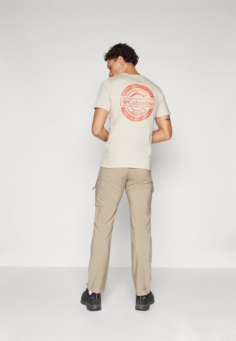 T-shirt en coton beige avec un logo circulaire rouge de Columbia au dos, associé à un pantalon cargo beige clair et des chaussures noires. Design simpliste.