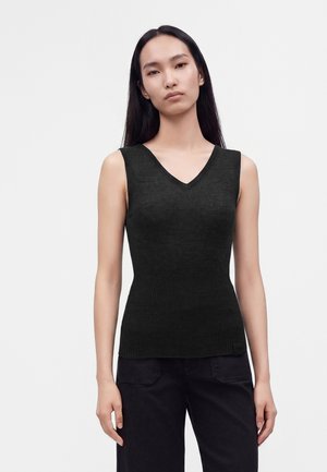 Donna con lunghi capelli scuri che indossa una maglietta aderente nera senza maniche con scollo a V e pantaloni neri su uno sfondo chiaro e semplice.