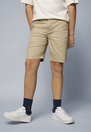 Beige Baumwollshorts mit geradem Schnitt, ausgestattet mit seitlichen Taschen. Getragen mit weißen Turnschuhen und dunkelblauen Socken, vor einem grauen Hintergrund stehend.