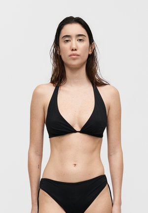 Γυναίκα με μακριά σκούρα μαλλιά που φοράει μαύρο μπικίνι με halter λουρί και μαύρο κάτω μέρος μαγιό, μπροστά από απλό φόντο.