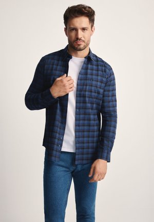 Niebiesko-czarna koszula w kratę wykonana z tkaniny, z długimi rękawami, zapinana na guziki z subtelnym detalem kieszeni, noszona na białym t-shircie i dżinsach.