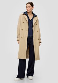 Beige dubbelzijdige trenchcoat met gouden knopen, gedragen over een marineblauwe hoodie en zwarte broek, aangevuld met witte schoenen.