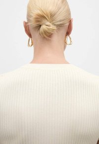 Blond vlasy stočené do nízkého drdolu, nosí nepravidelné zlaté kruhové náušnice a krémový žebrovaný top, zobrazená zezadu na bílém pozadí.