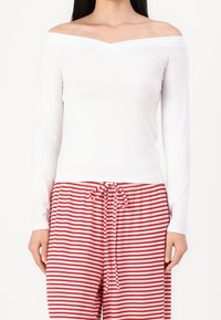 Bílý off-shoulder top s dlouhým rukávem zkombinovaný s červenými a bílými pruhovanými pyžamovými kalhotami se stahovací šňůrkou v pase. Hladký materiál a přiléhavý design.