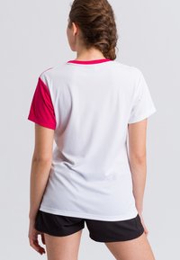 Maglietta sportiva bianca con accenti rosa sulle spalle, maniche corte e tessuto texturizzato. Abbinata a pantaloni corti neri, vista da dietro.