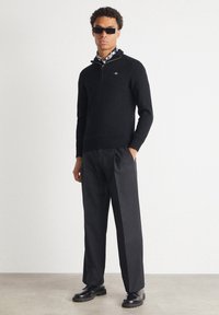 Hombre de pie con gafas de sol negras, suéter negro con media cremallera, bufanda estampada, pantalones negros y zapatos de cuero negros sobre un fondo liso.