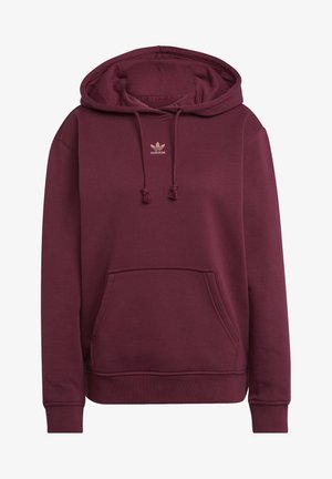 Henkilö pukeutuneena viininpunaiseen Adidas-huppariin, jossa on etutasku ja kiristysnauhat, yhdistettynä luonnonvalkoisiin housuihin, jotka on lyhennetty kaulan ja reisien kohdalta.