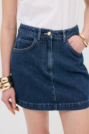 Minifalda de mezclilla en azul oscuro, con cintura fruncida, bolsillos frontales y cierre de botón dorado. Textura suave con costuras en contraste.