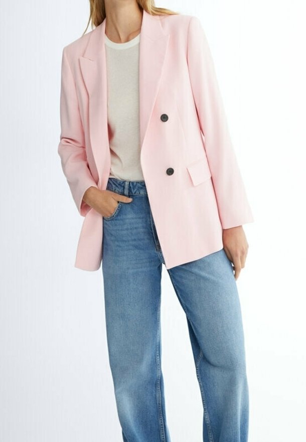 Femme portant un blazer croisé rose clair sur une chemise blanche, associé à un jean taille haute bleu, debout avec une main dans la poche.